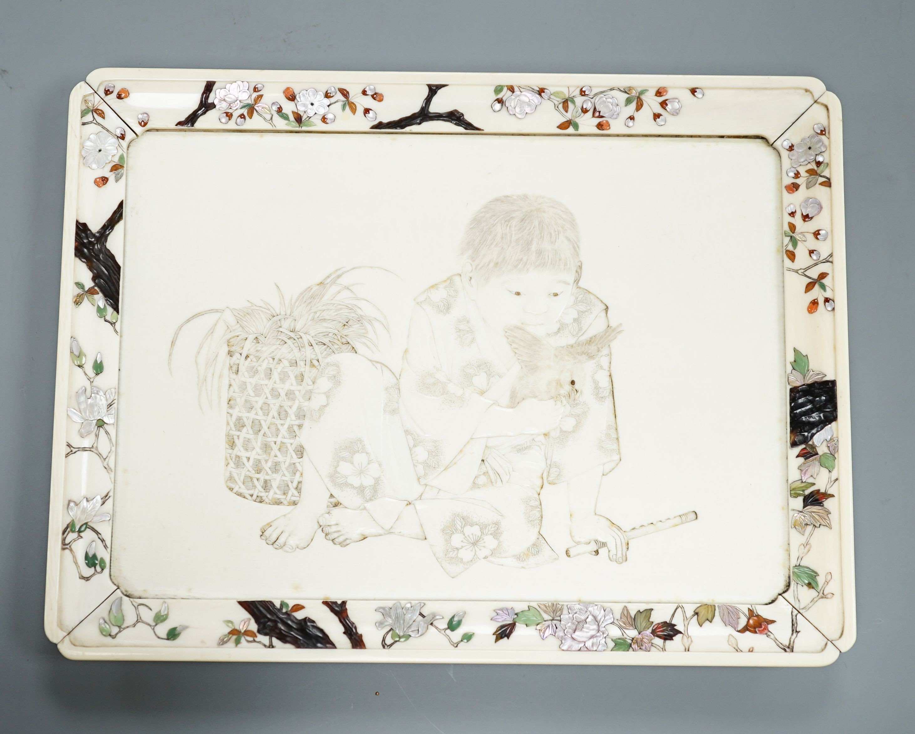 A Japanese ivory tray/plaque, Meiji period, with sunk-in carving of boy with dove, the surround decorated with Shibayama inlaid flowers etc. 15 x 20cm